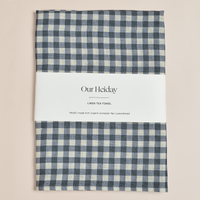 A folded linen tea towel with a blue and cream gingham check pattern, wrapped in a white paper band labeled 'Our Heiday LINEN TEA TOWEL'. The towel is photographed flat against a solid, warm beige background.