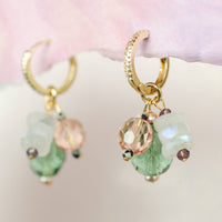 Gold hoop earrings with green and pink gemstones on a soft pink background