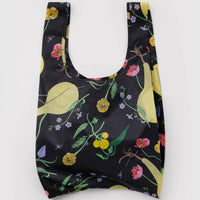 A reusable shopping bag with a black background and a colorful botanical print featuring yellow pears, green leaves, pink flowers, and small purple blossoms, shown against a plain light gray background.