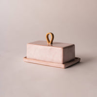 A rectangular pink butter dish with a matching lid and small loop handle, shown on a plain warm beige studio background.