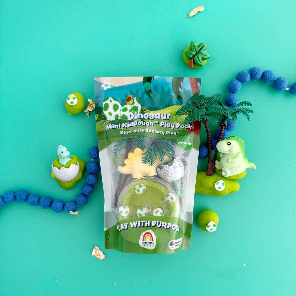 A package of Mini Dinosaur Play Puddle sensory play sand in a clear bag with a green label, displayed on a bright turquoise background with small dinosaur toys and green palm decorations around it.