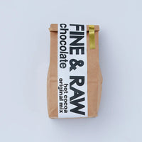 A brown kraft paper bag of Fine & Raw Hot Cocoa Original Mix, featuring a white label with bold black text and a gold ribbon accent, set against a clean light gray background.