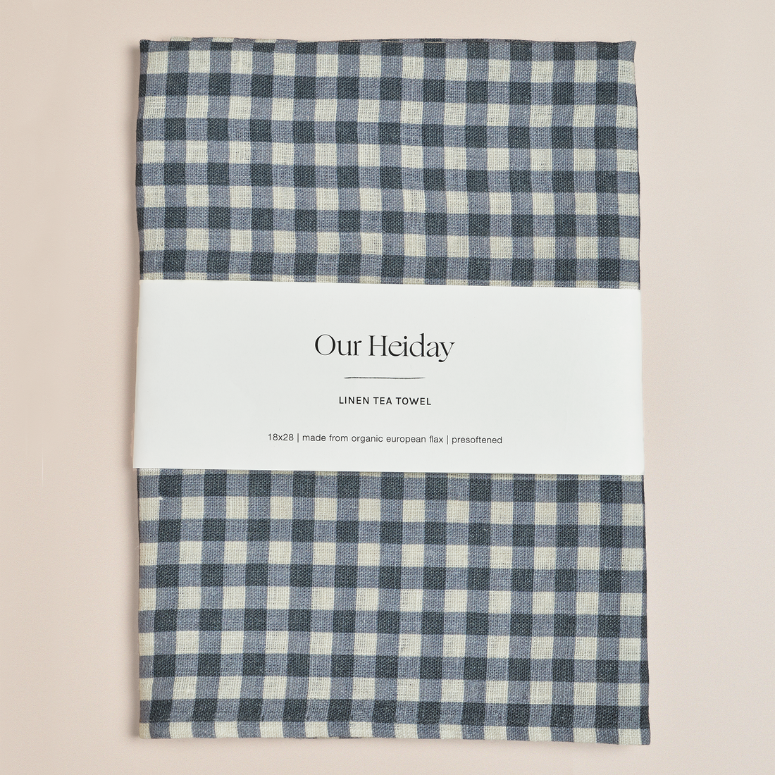 A folded linen tea towel with a blue and cream gingham check pattern, wrapped in a white paper band labeled 'Our Heiday LINEN TEA TOWEL'. The towel is photographed flat against a solid, warm beige background.