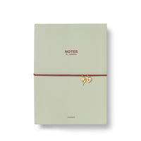 A sage green soft PU cover notebook with a red elastic band closure adorned with two small gold charms A5