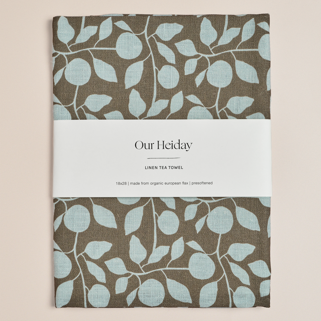 A folded linen tea towel with a brown and light blue leaf pattern, wrapped in a white paper band labeled 'Our Heiday'.
