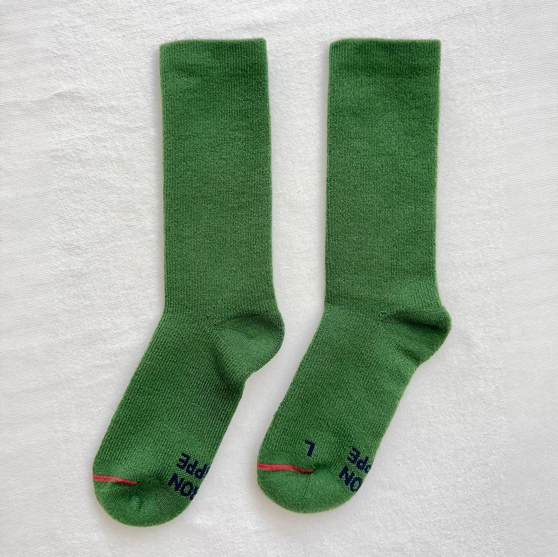A pair of green crew socks laid flat on a white textured background, with dark navy text and a thin red stripe near the toes.