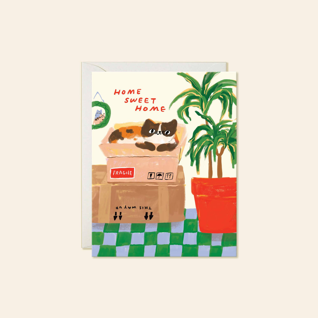 A greeting card with a whimsical illustration of a calico cat resting in a cardboard shipping box next to a potted palm tree, with the text 'HOME SWEET HOME' 