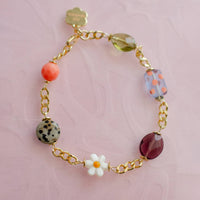 A gold chain bracelet with various colorful beads, including a white daisy charm, a speckled green bead, a coral orange bead, and a purple teardrop bead. 