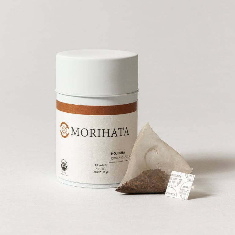 A white cylindrical canister of MORIHATA organic hojicha tea with a brown stripe and logo, shown next to a single triangular paper tea bag containing dark brown tea powder on a light gray background.