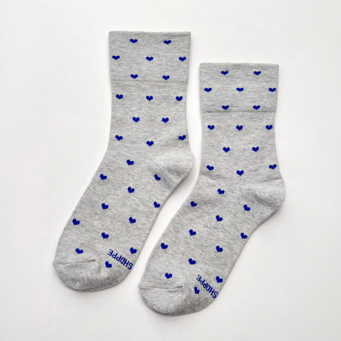 A pair of light heather gray crew socks with a repeating pattern of small blue hearts, featuring the brand name 'SHOPIE' printed in blue near the toes.